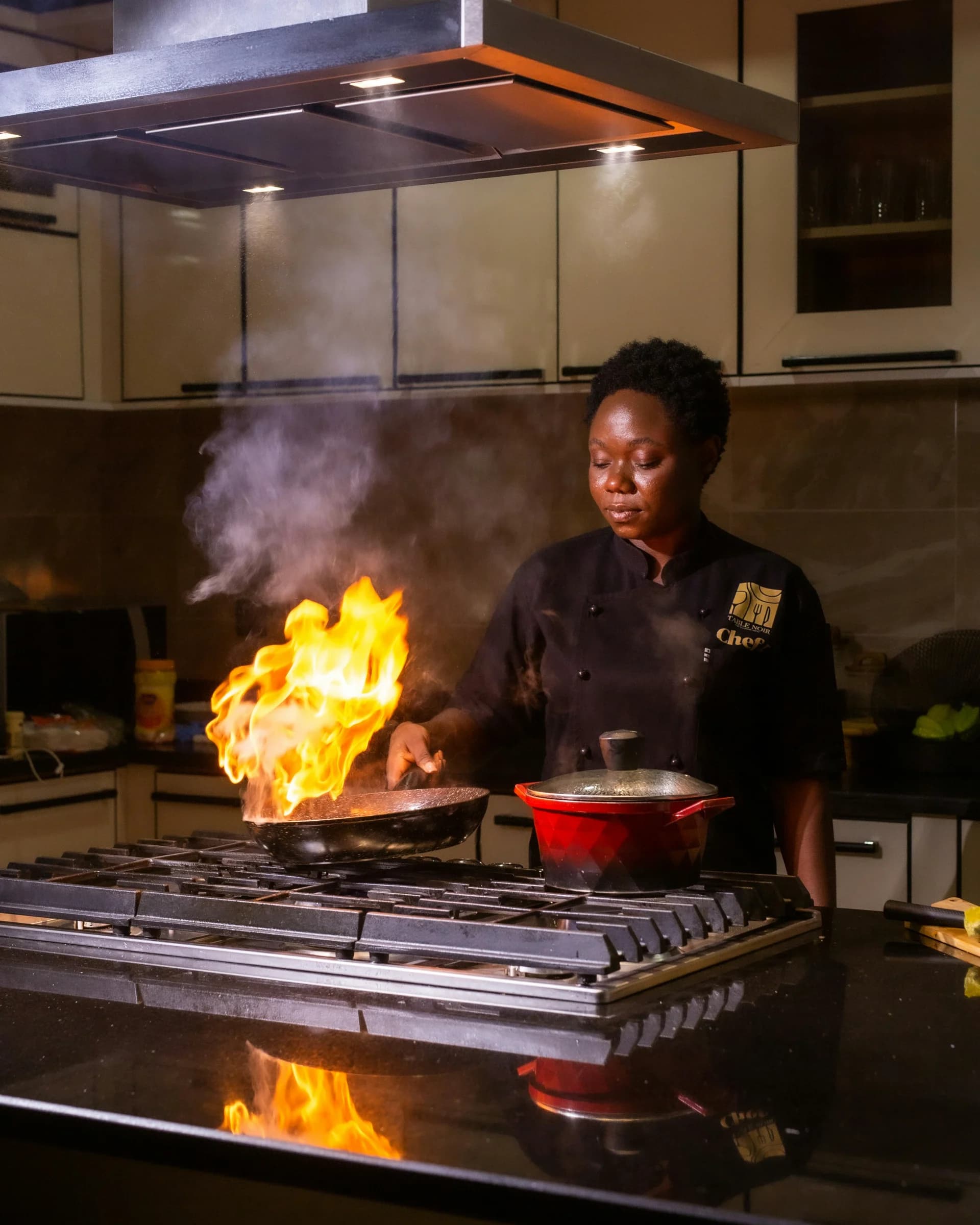 Une cheffe en uniforme noir fait flamber des aliments dans une poêle sur une cuisinière à gaz, avec une cocotte rouge à côté.